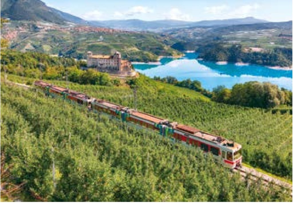 Treno della Trento-Malè tra i meleti sopra il lago di Santa Giustina, con un castello sullo sfondo